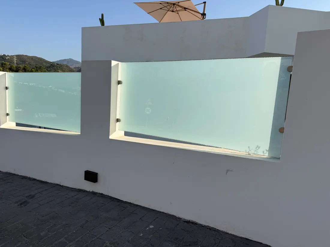 Muro perimetral blanco con paños de vidrio translúcido fijados con botones, visto desde la parcela