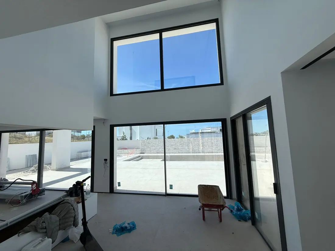 Interior a doble altura en obra con ventanales y correderas de aluminio negro y suelo protegido